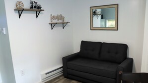 A room with a black sofa, wooden shelves with decorative items, a wall-mounted mirror, and a white wall with a power outlet.
