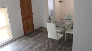 A dining area with a glass table and white chairs, a wooden door, and a view into a bedroom.