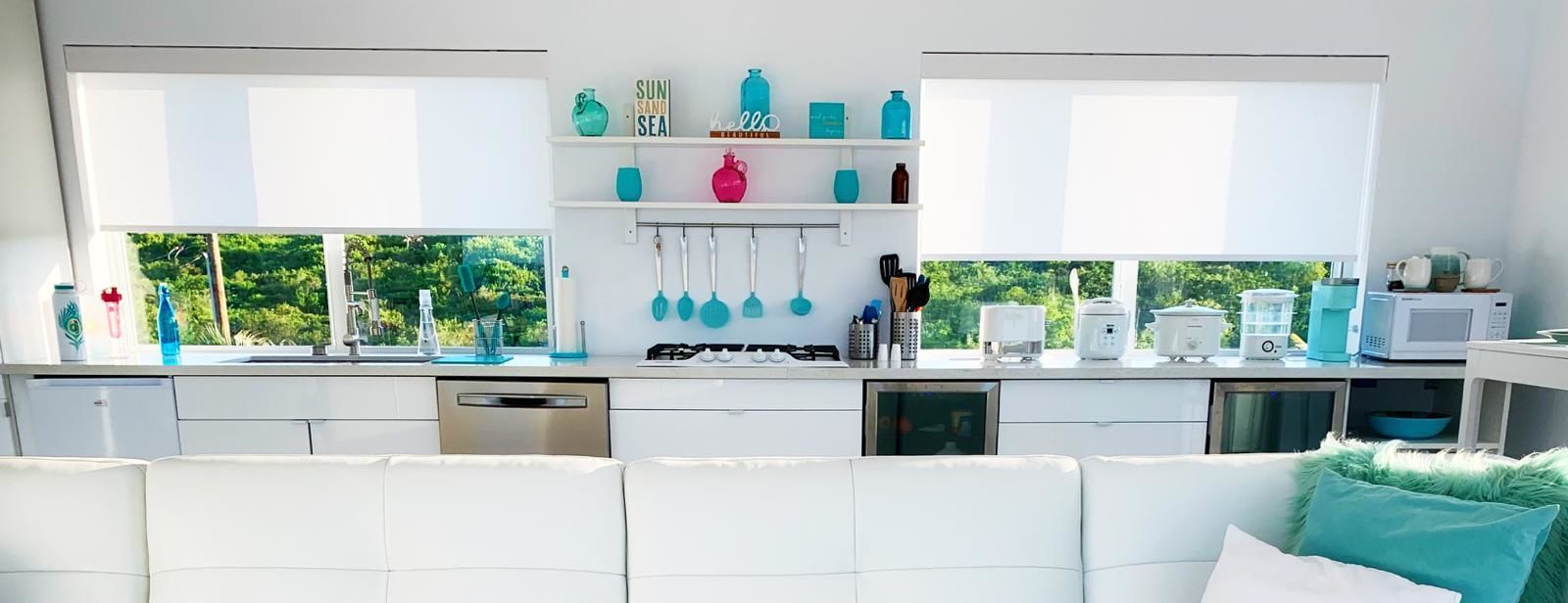 Una cocina moderna con armarios blancos, estufa a gas, microondas y cafetera. Se observan repisas con objetos decorativos y utensilios colgando sobre la encimera.