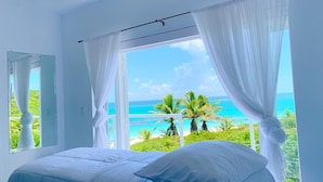 A bedroom with a bed, white curtains, and a view of the ocean and palm trees. - Mika's Resort (Governor's Harbour)