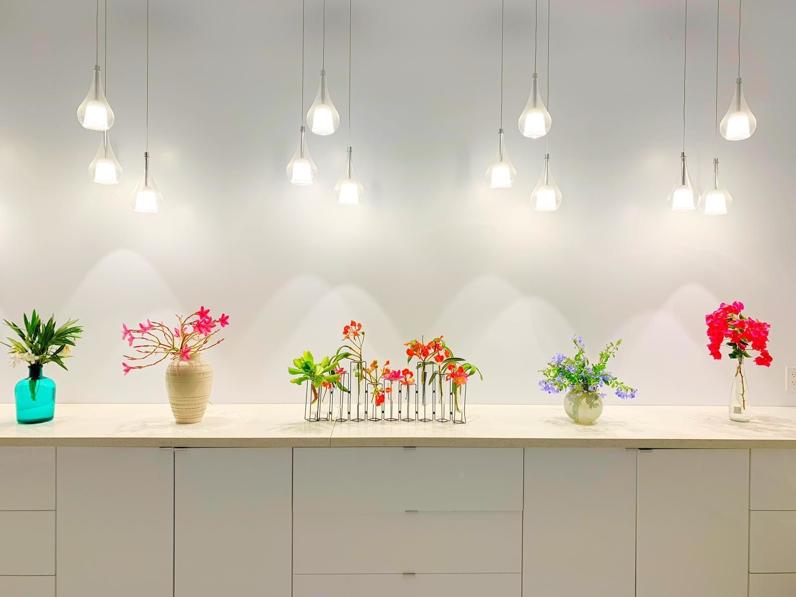 A kitchen counter with multiple hanging light fixtures and various flower arrangements.