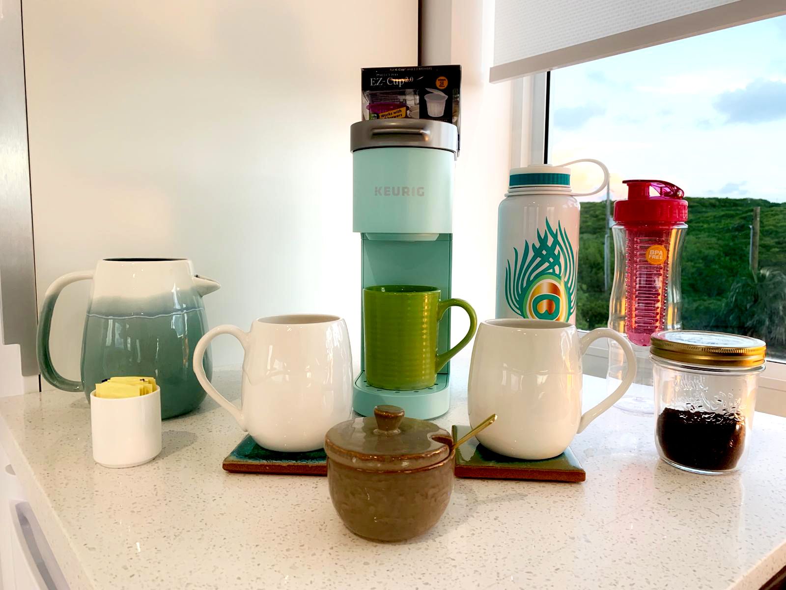 Un comptoir de cuisine avec divers mugs à café et à thé, un distributeur d’eau, une théière et un bocal de café.