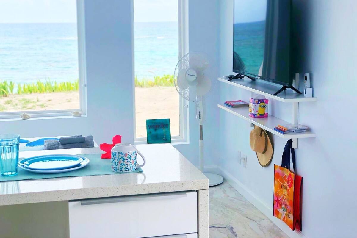 Ein Zimmer mit Fernseher auf einem weißen Regal, einem Ventilator, einem Fenster mit Blick auf den Strand und einem Schreibtisch mit Tasse und Tellern.