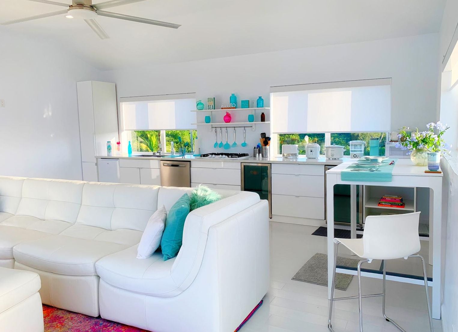 A modern kitchen with white cabinetry, a dining area with a white table and chairs, and a white sofa with teal cushions.
