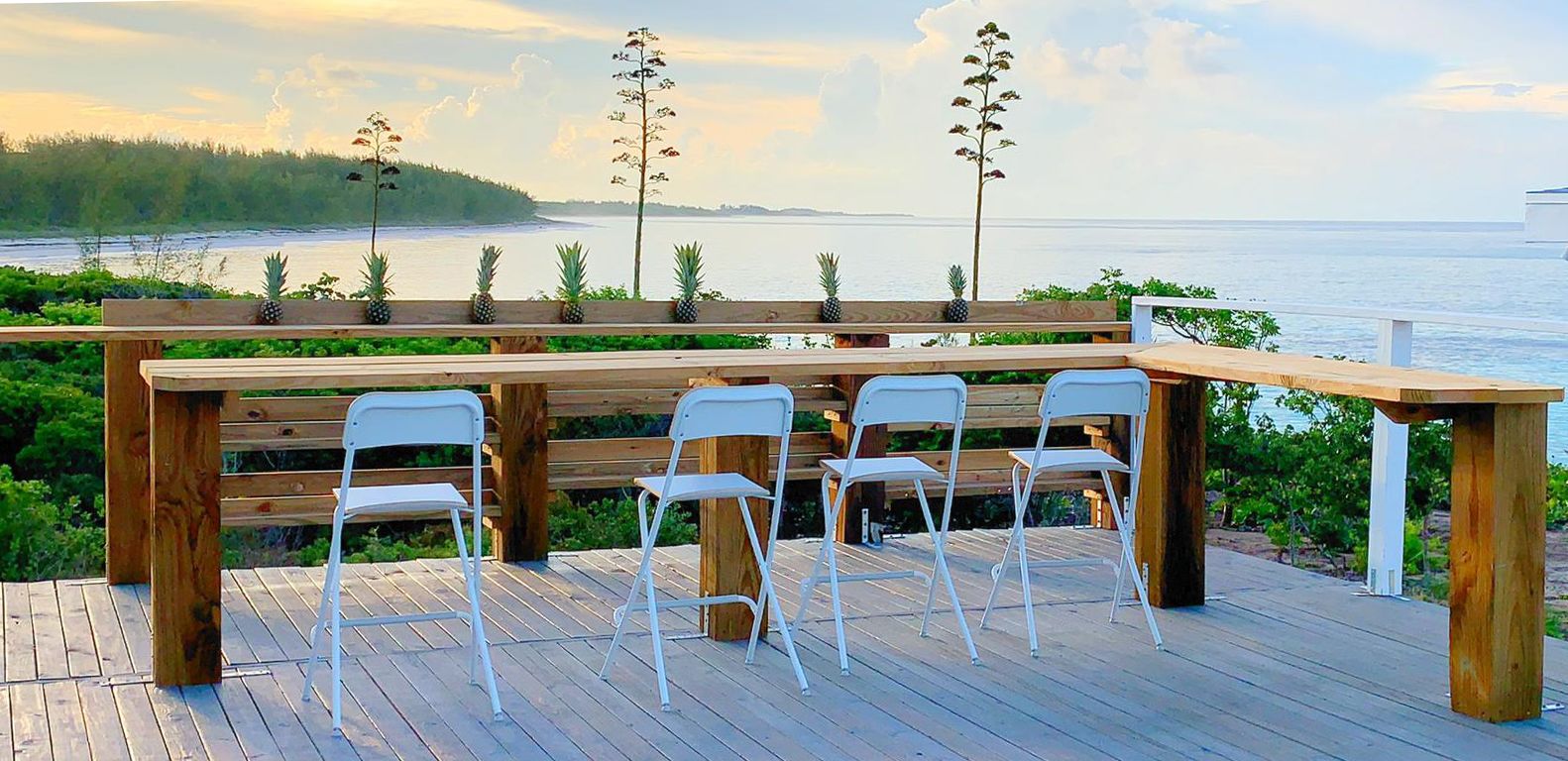 A wooden deck with a table and four white chairs, overlooking a body of water and a forested area.