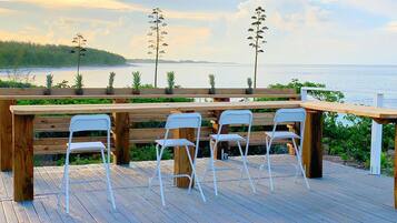 A wooden deck with a table and four white chairs, overlooking a body of water and a forested area.