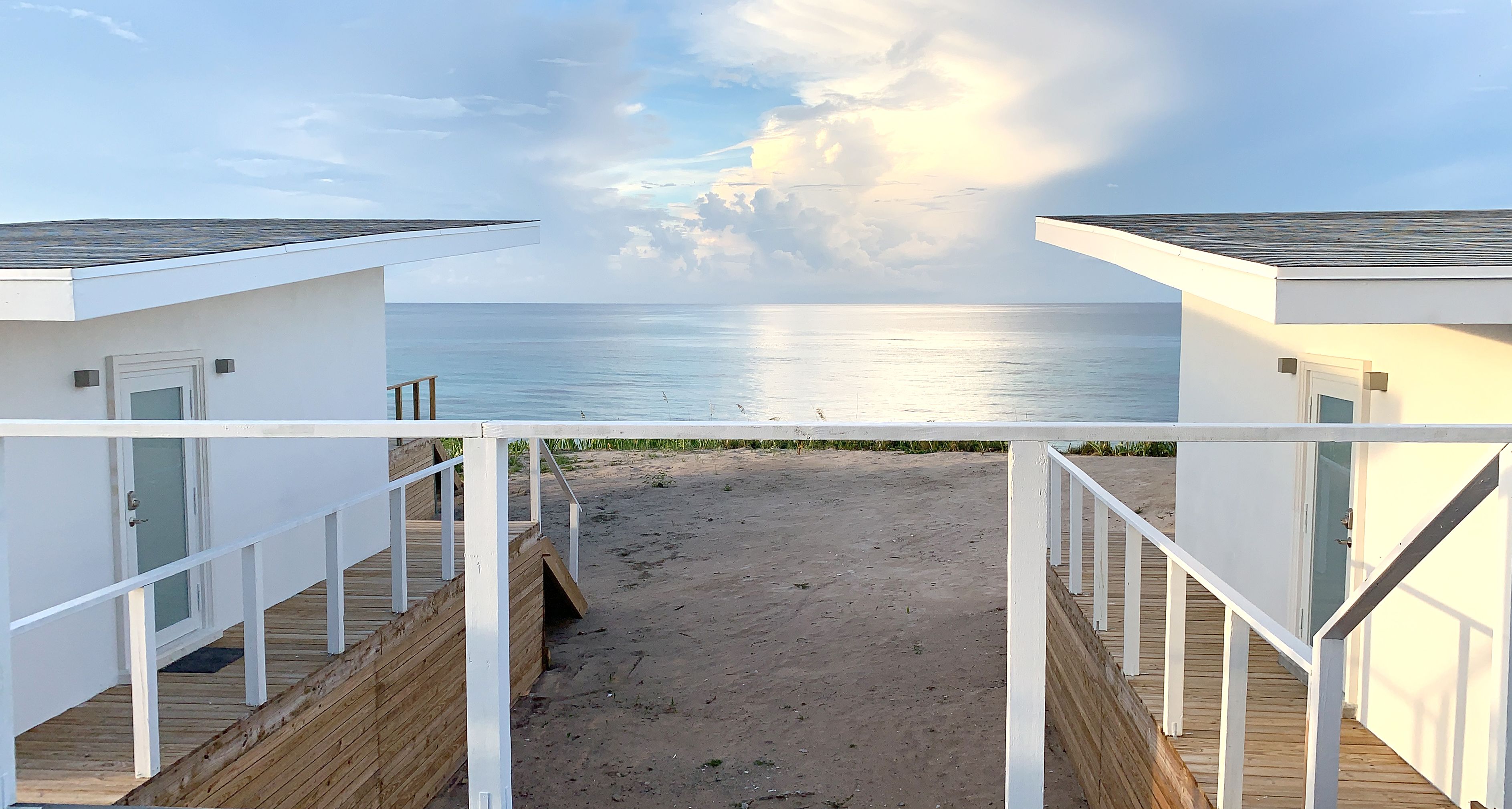 A beachside view with a wooden deck leading to a sandy beach, white buildings with doors, and a clear view of the ocean.