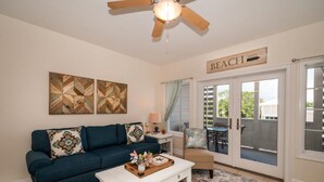 Una sala de estar con un sofá azul oscuro, una mesa de centro blanca con una bandeja, y un ventilador de techo. Un letrero en la pared dice "BEACH 500 FT".