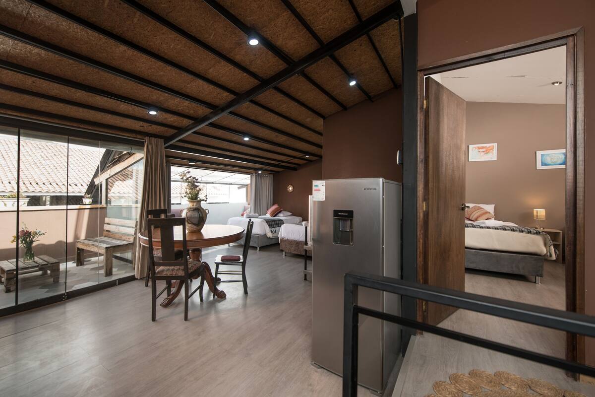 A modern hotel room with a wooden ceiling, a dining area with a table and chairs, a bed with a nightstand, and a refrigerator.