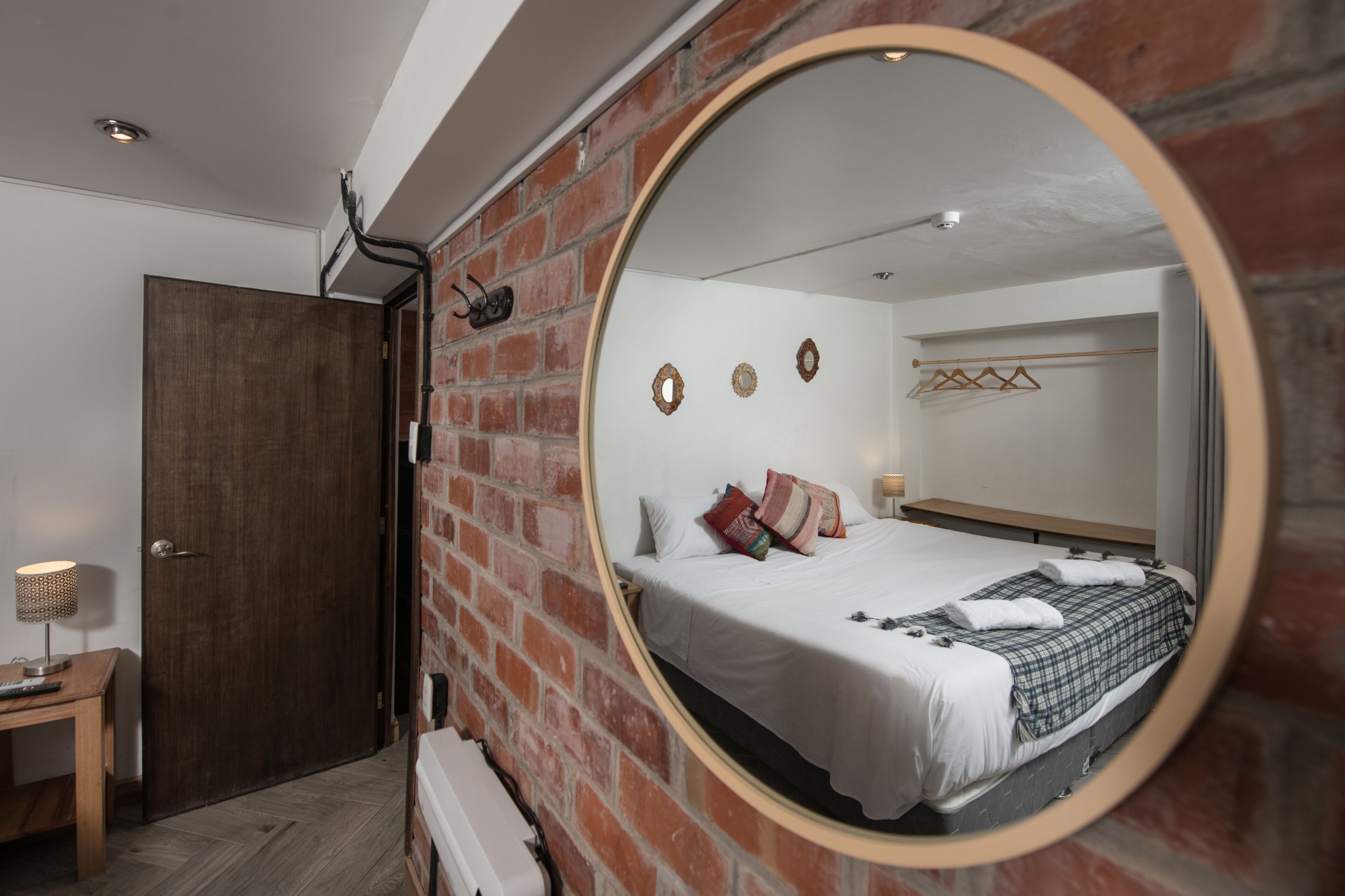 A bedroom with a brick wall, a round mirror, a bed with white bedding, a wooden nightstand, and a hanging light fixture.