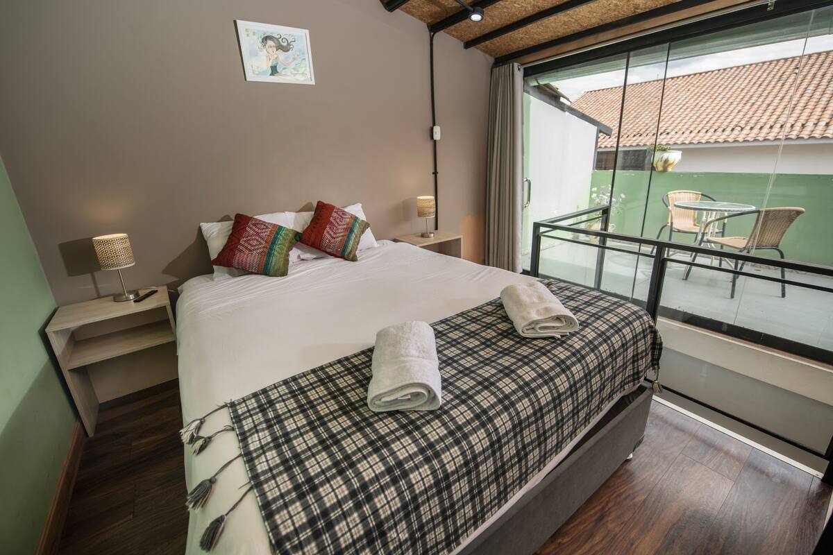 A hotel room with a bed, a nightstand, a lamp, a window with a view of the outside, and a balcony with chairs.