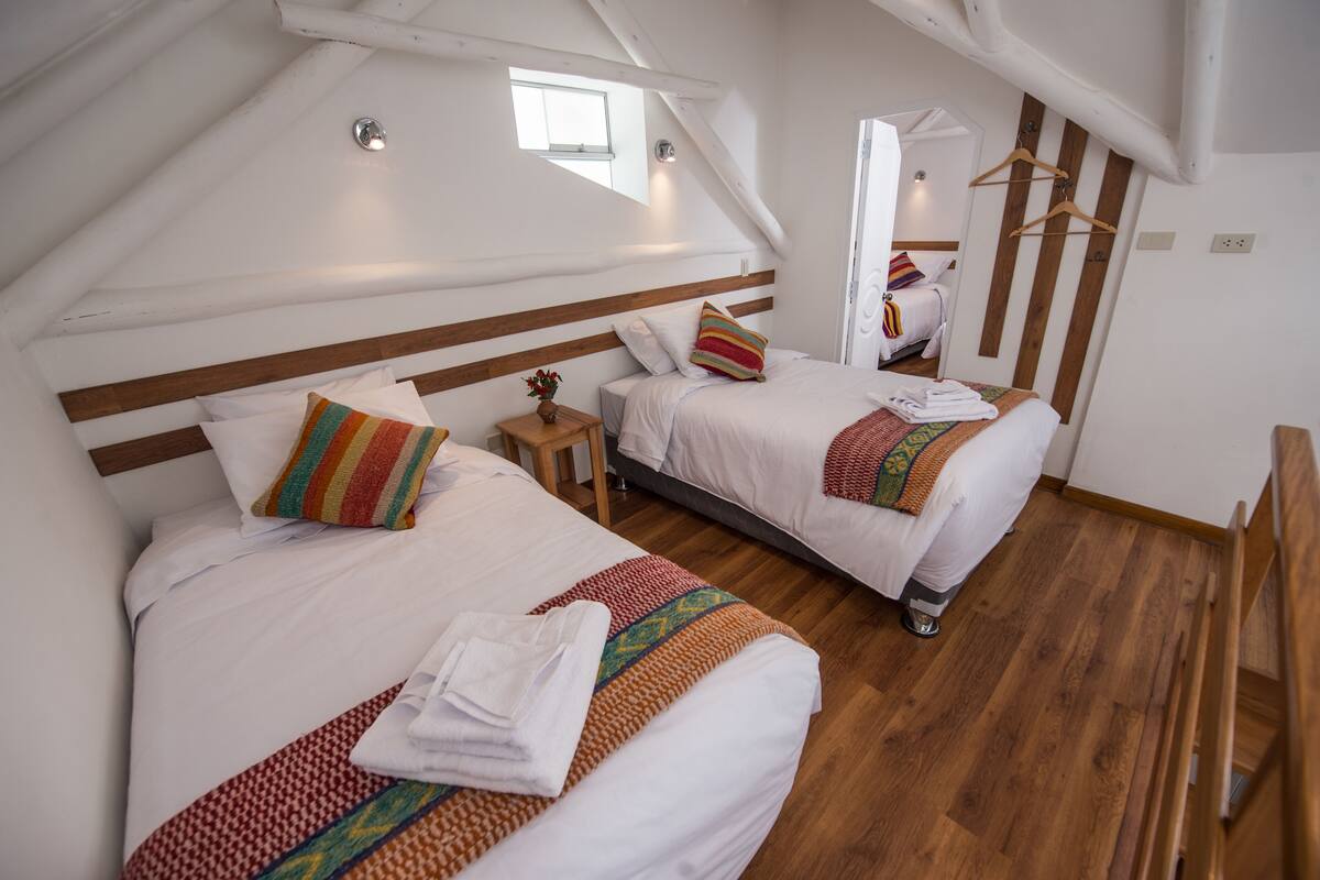 A bedroom with two beds, wooden headboards, a small bedside table with a lamp, and a window.