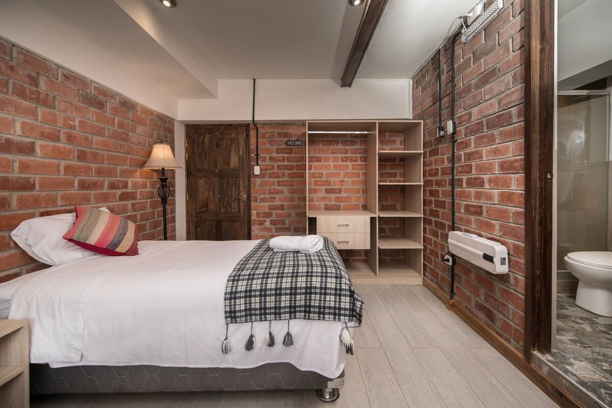 A bedroom with a brick wall, a bed with a plaid blanket, a wooden wardrobe, and a toilet.