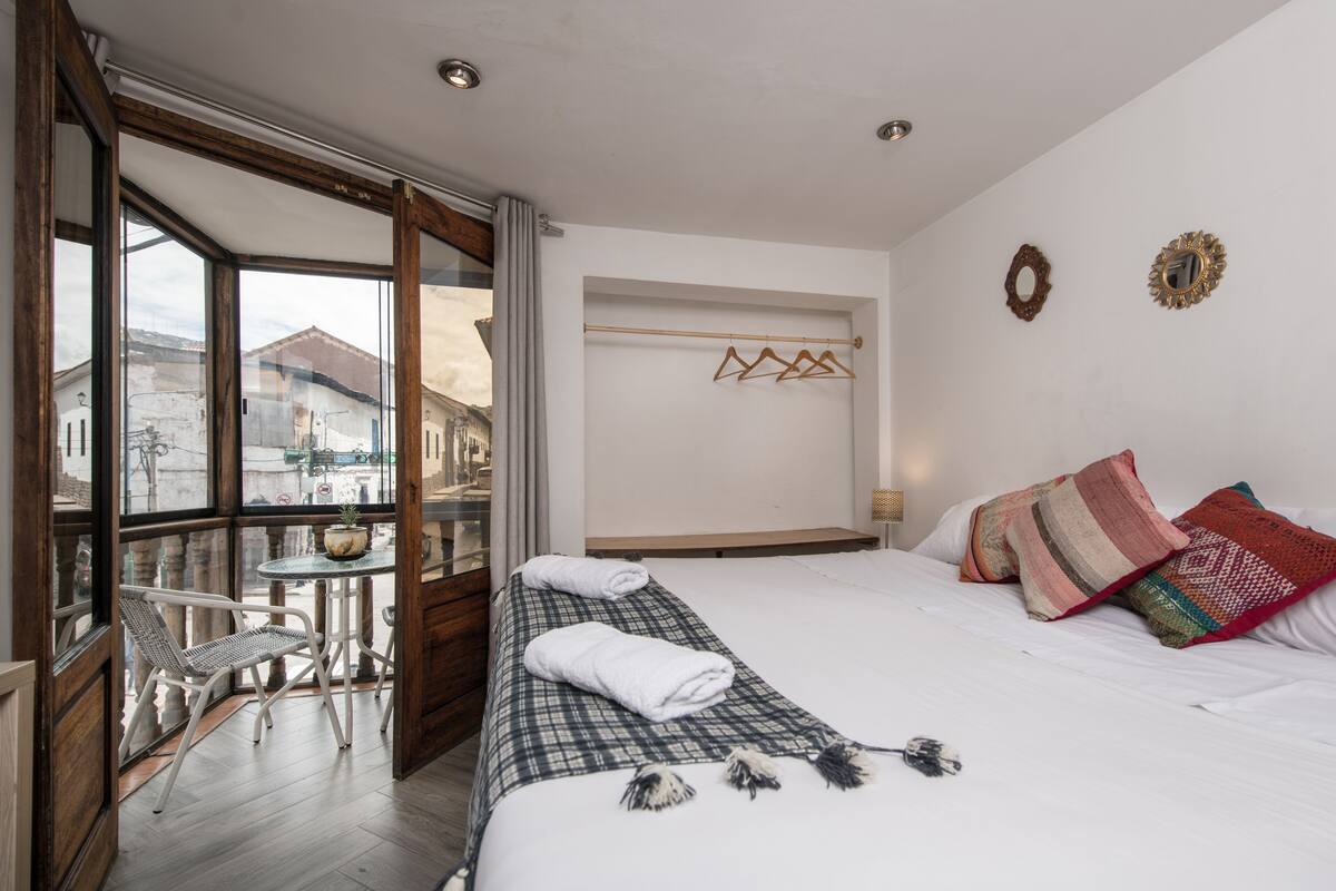 A hotel room with a bed, a small table with a chair, a window with a view of buildings, and a coat rack with hangers.