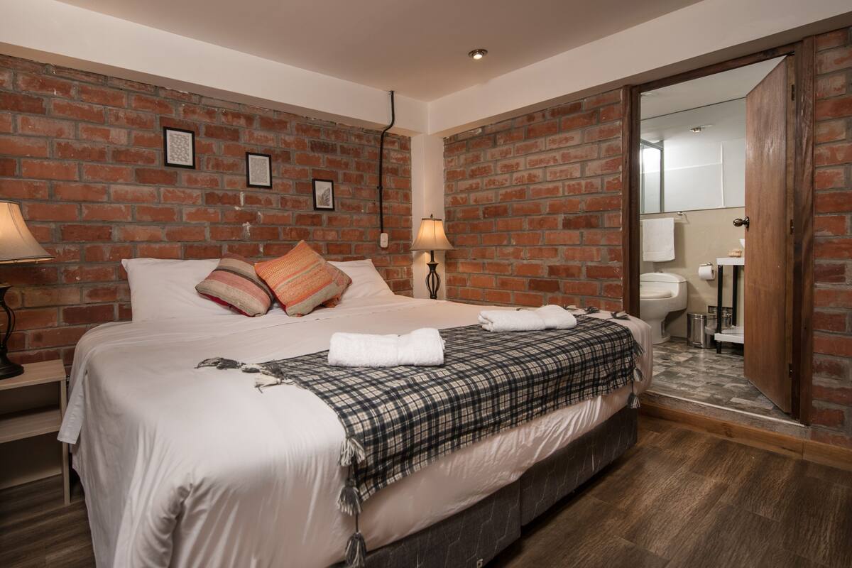 A hotel room with a brick wall, a large bed with white bedding, a nightstand, a lamp, and a bathroom visible through an open door.