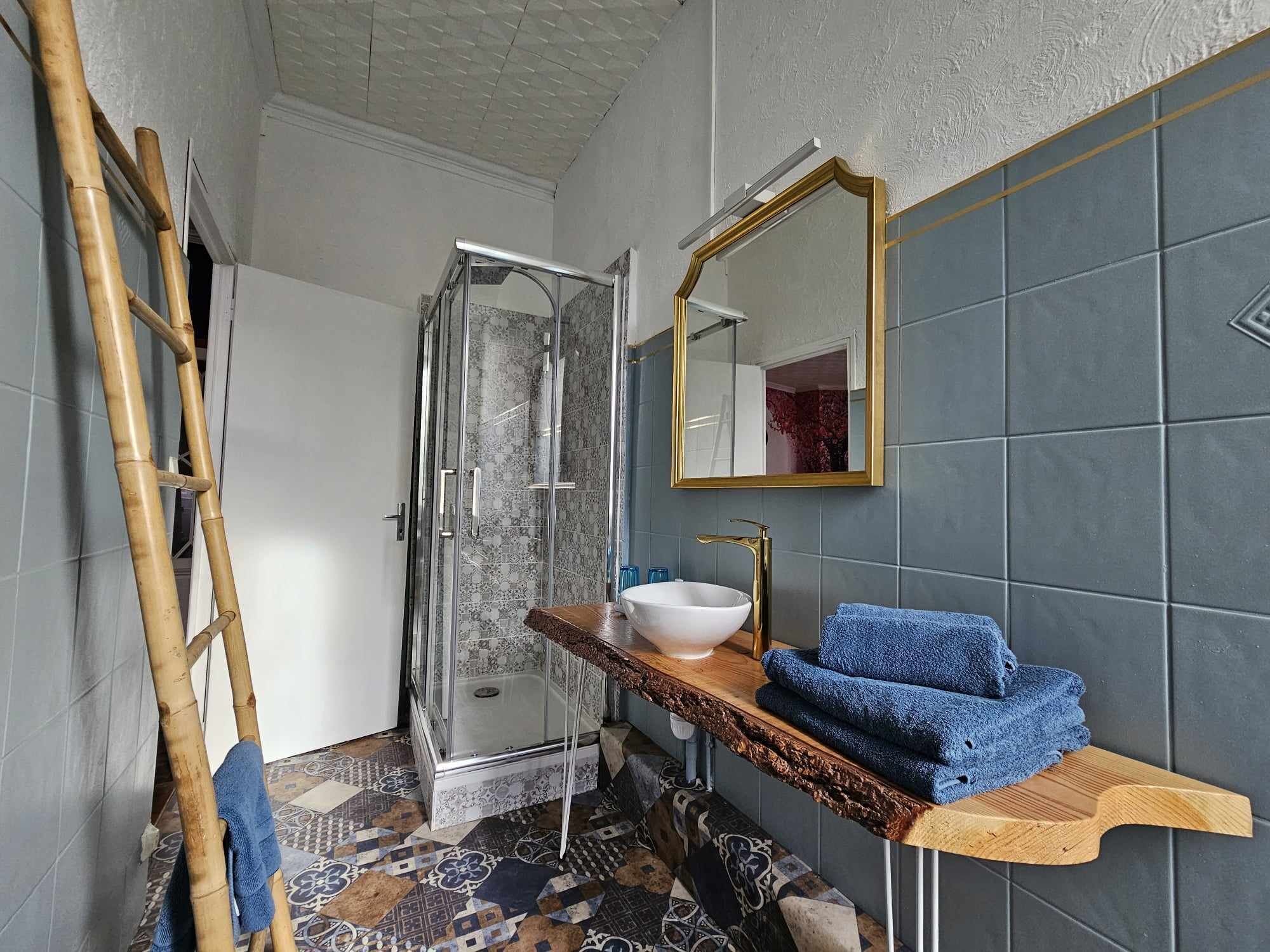 A bathroom with a glass shower enclosure, a white sink with a gold faucet, and a wooden vanity with blue towels.