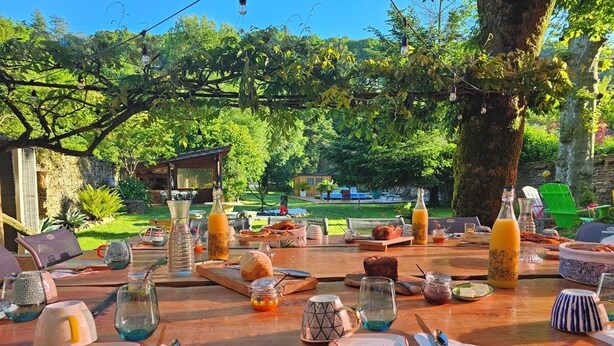 An outdoor dining area with a wooden table set for breakfast, surrounded by greenery and a view of a garden.