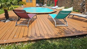 A wooden deck with a pool, surrounded by a stone wall and a small building with a tiled roof.