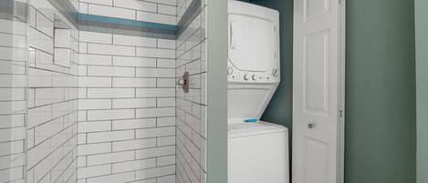 Un baño con paredes de azulejos blancos, zona de ducha y una lavadora blanca.