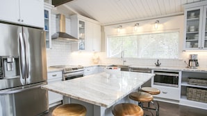 Une cuisine moderne avec des meubles blancs, des appareils en acier inoxydable, un îlot en marbre avec des tabourets et une grande fenêtre avec des stores.