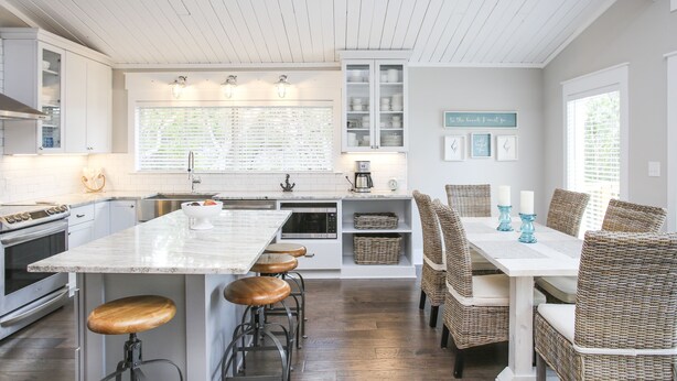 Une cuisine moderne avec des meubles blancs, un grand îlot central doté de tabourets et un coin repas avec des chaises en osier.