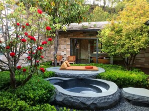 A modern outdoor seating area with a stone fire pit, lush greenery, and a building with a glass door in the background.