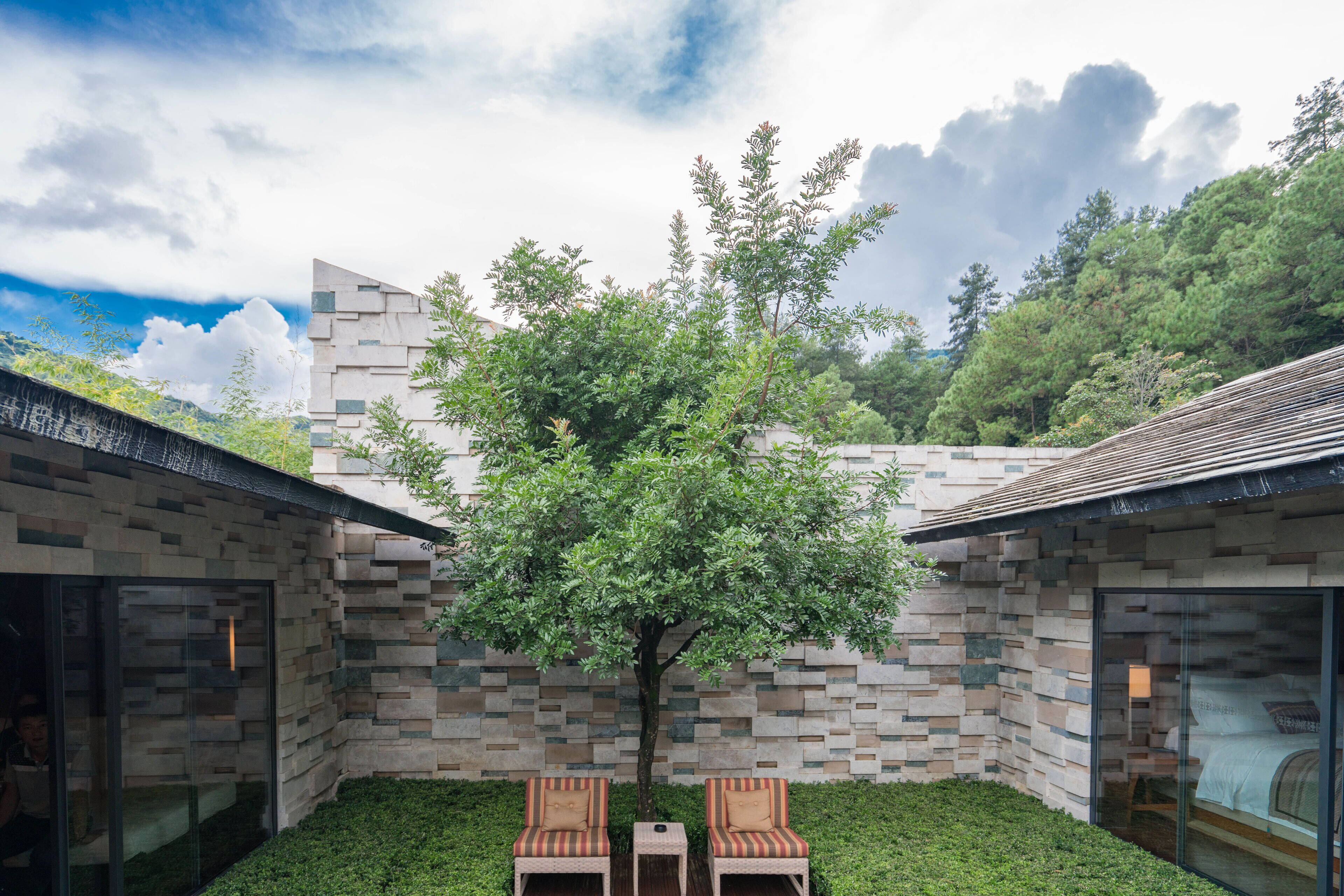 A modern building with a stone facade, two outdoor seating areas, and a tree in the center.