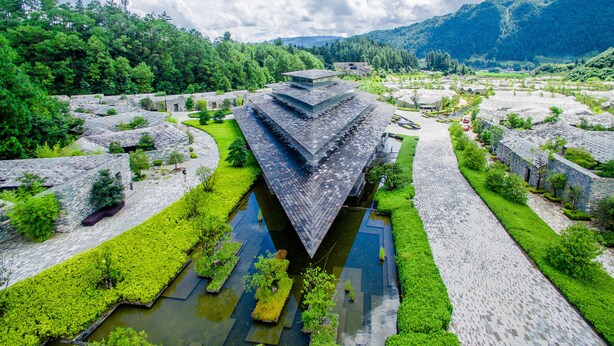 Un complejo arquitectónico tradicional con tejados escalonados, rodeado de abundante vegetación y un pequeño elemento acuático.