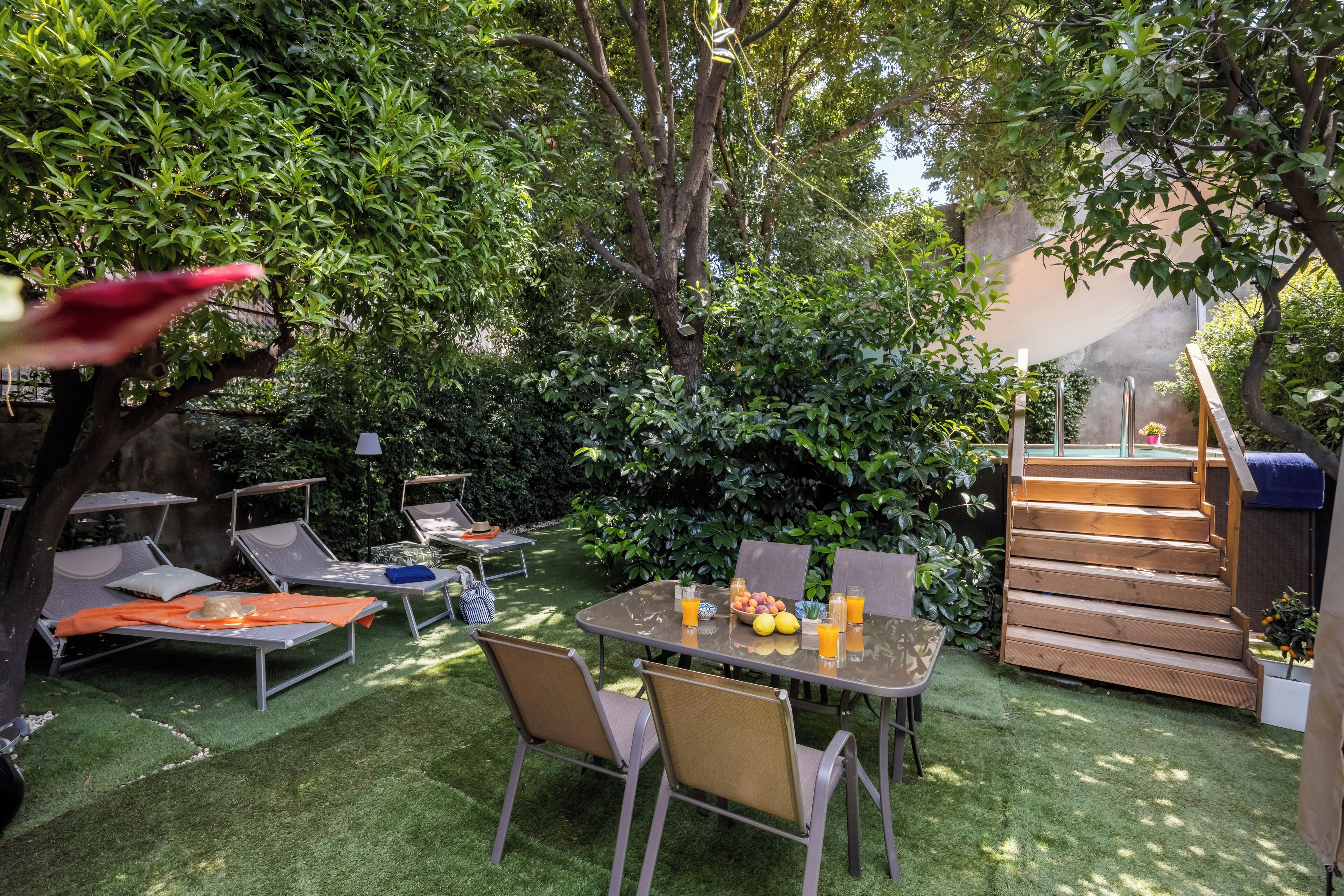 Une terrasse avec une table dressée pour deux personnes, entourée de chaises longues, d’un escalier en bois et d’une végétation luxuriante.