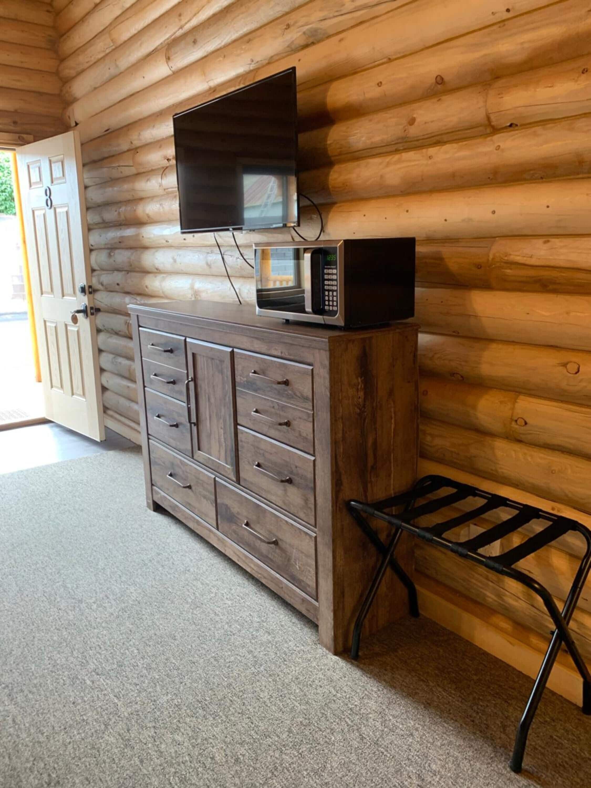 A wooden cabin interior with a wooden cabinet, a microwave, a television mounted on the wall, and a small stool.