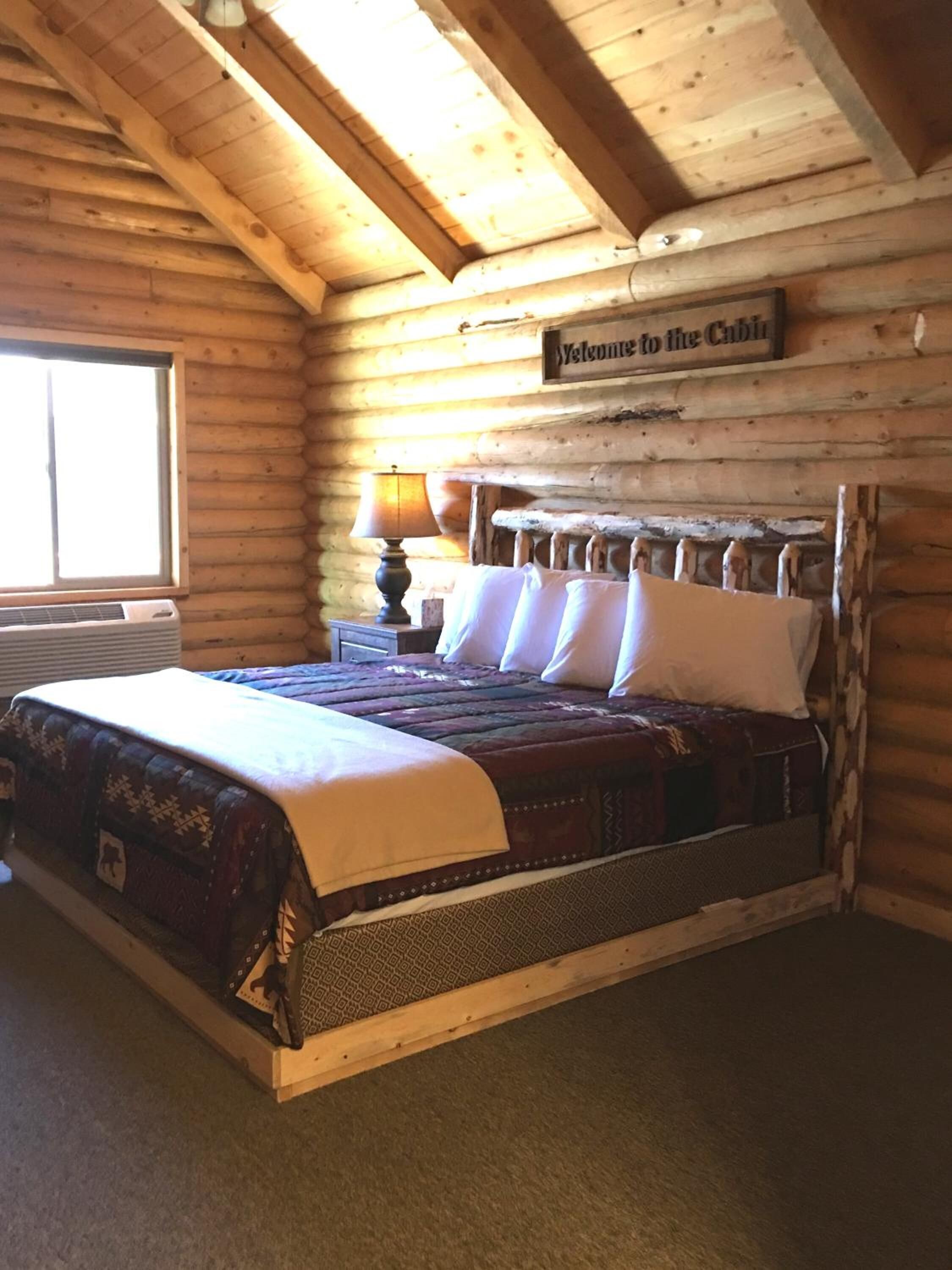 A log cabin bedroom with a bed, a nightstand, a lamp, and a 'Welcome to the Cabin' sign.