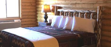 A log cabin bedroom with a bed, a nightstand, a lamp, and a 'Welcome to the Cabin' sign.