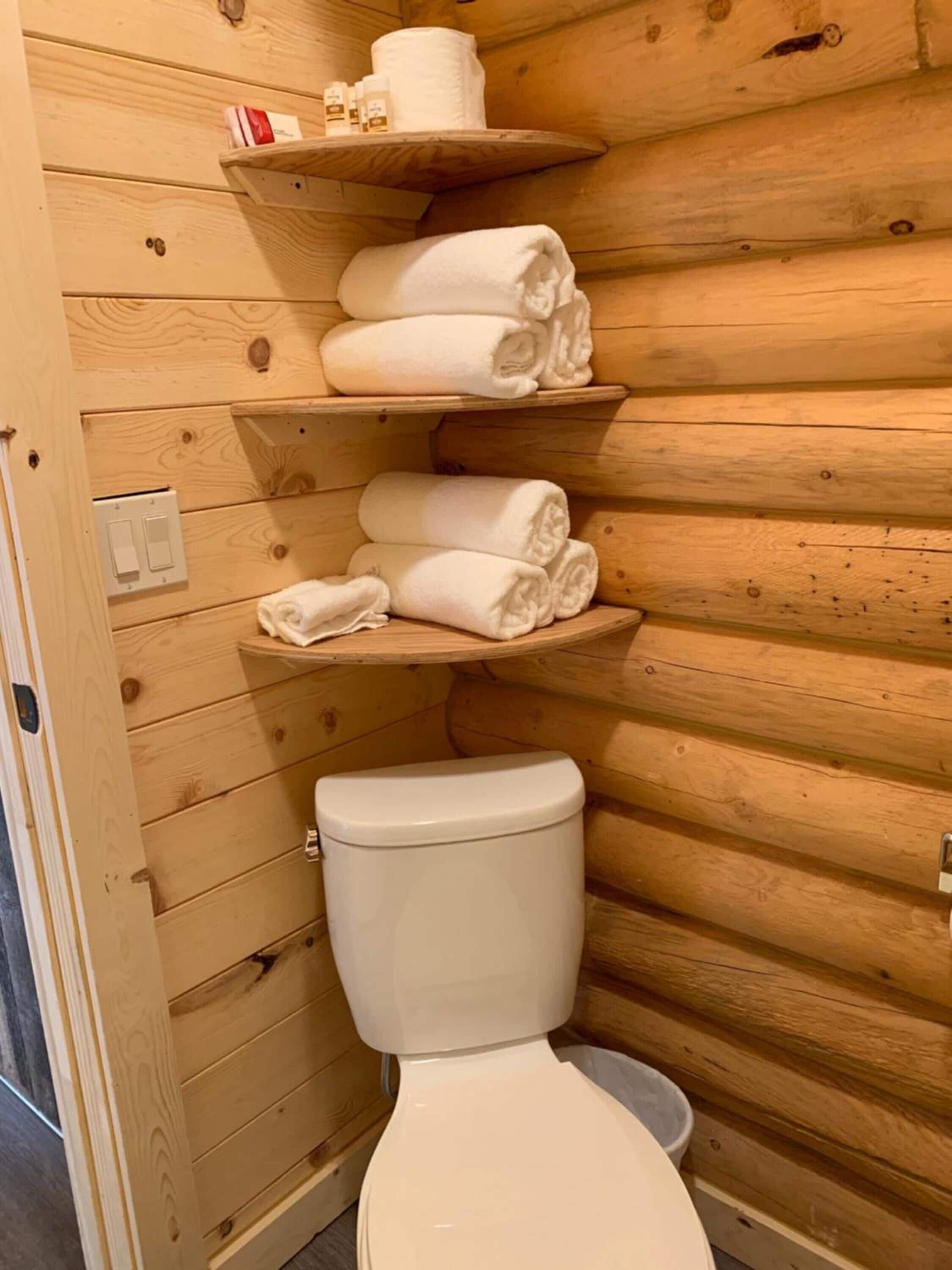 A wooden cabin bathroom with a toilet, shelves holding towels, and toiletries.