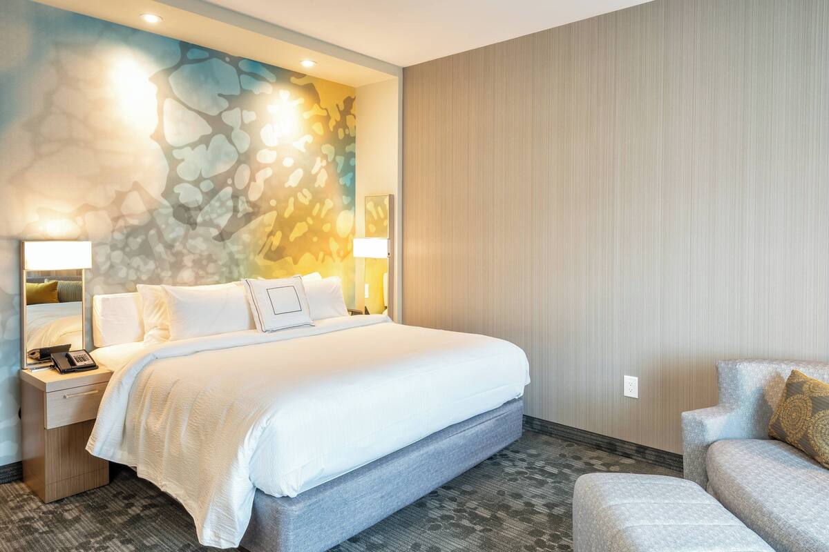 Ein Hotelzimmer mit einem großen Bett, einem Nachttisch mit Lampe, einer kleinen Sitzecke und einer dekorativen Wandmalerei.