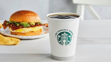 Een witte beker met een Starbucks logo en tekst, een hamburger en een gele servet op een witte tafel.