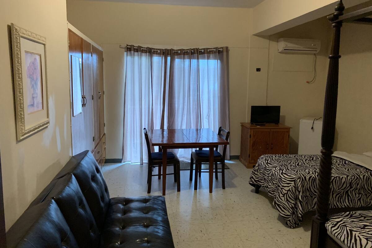 Una habitación con un sofá de cuero negro, una mesa de comedor de madera con sillas, una cama con estampado de cebra y un televisor sobre un mueble de madera.