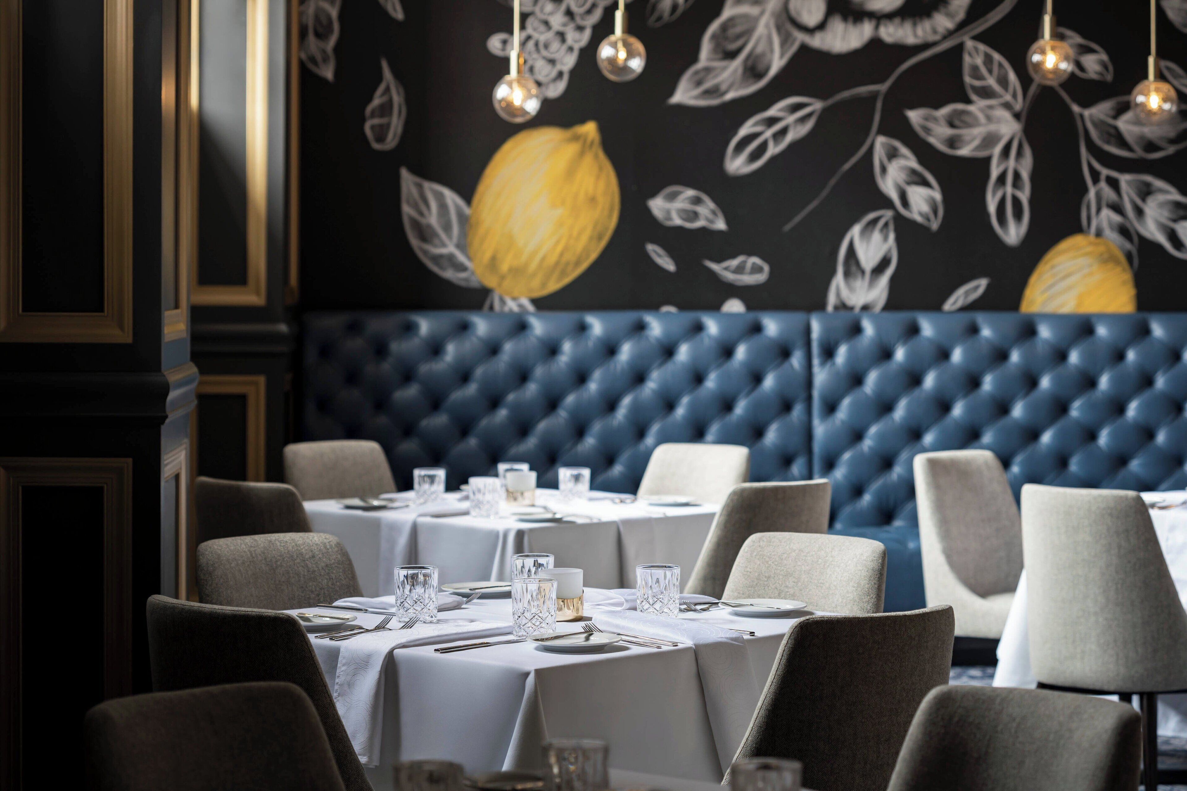 A restaurant with tables set for dining, a blue upholstered wall, and a wall mural with a lemon.