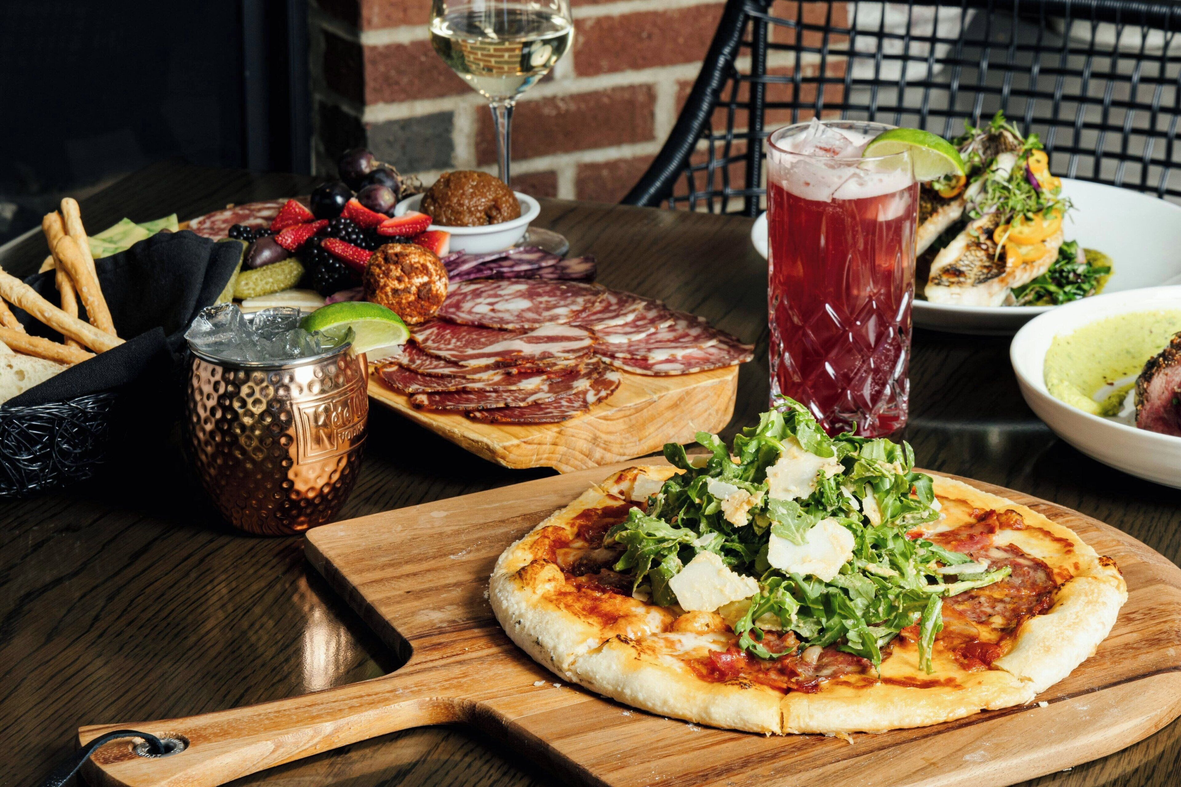 Una mesa de madera con pizza, un vaso de bebida roja, un bol de ensalada y un plato con carnes y quesos variados.