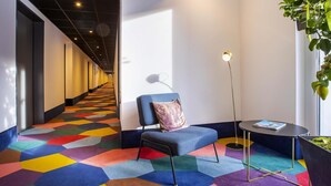 A hallway with a colorful geometric carpet, a blue sofa with a cushion, a small table with a magazine, and a tall floor lamp. - ibis Styles Pforzheim (Pforzheim)