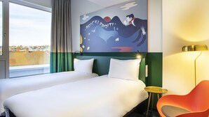 A hotel room with a large bed, a view of the city, a colorful painting on the wall, and a modern chair. - ibis Styles Pforzheim (Pforzheim)