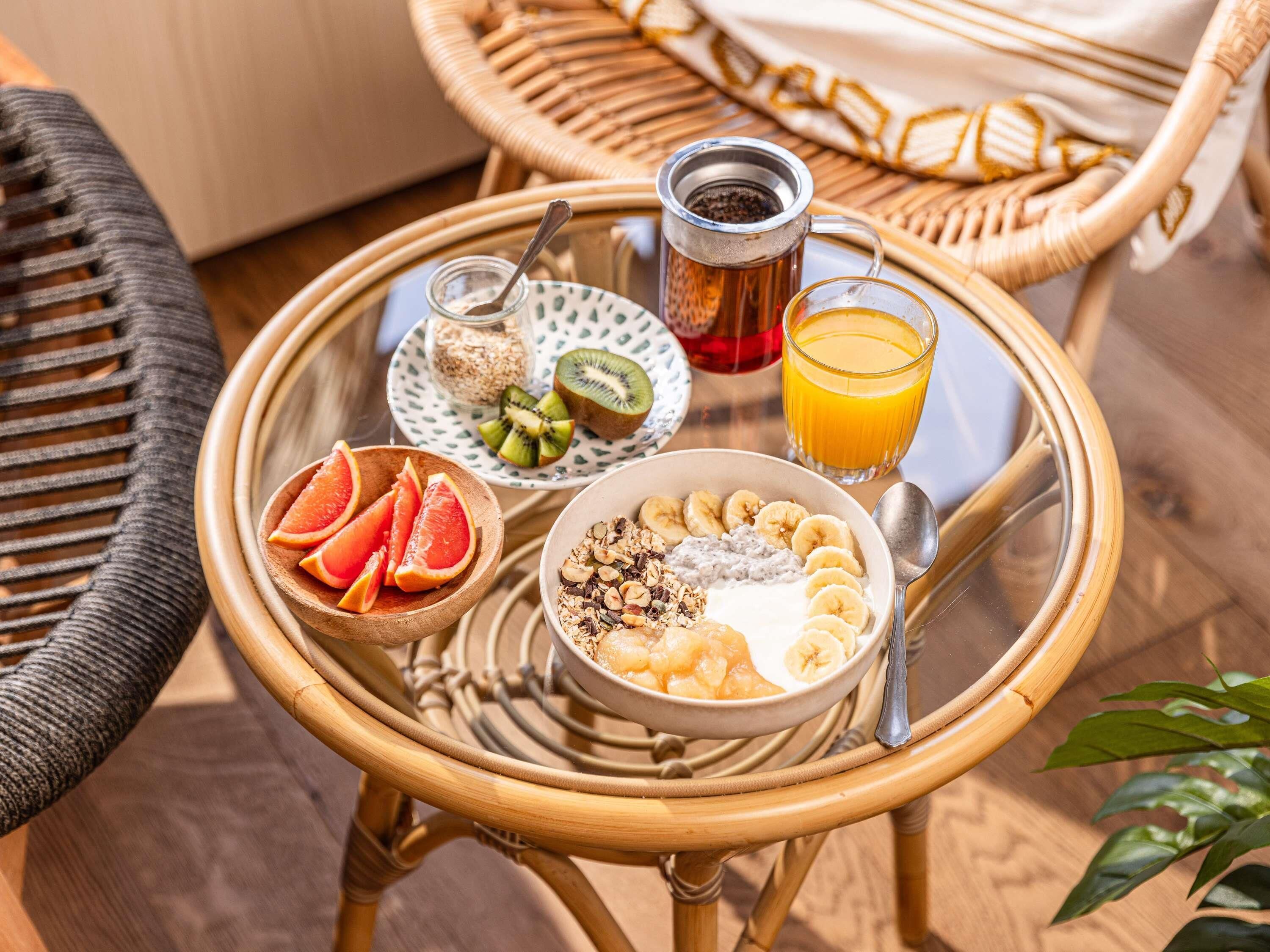 Ein Frühstücksbrett mit einer Schüssel Müsli, Bananenscheiben, Kiwis, Grapefruit, einem Glas Orangensaft, einer Tasse Tee und einem kleinen Honigglas.
