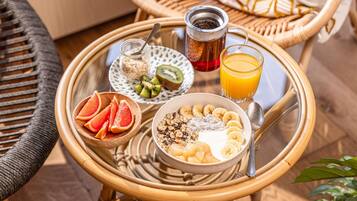 Ein Frühstücksbrett mit einer Schüssel Müsli, Bananenscheiben, Kiwis, Grapefruit, einem Glas Orangensaft, einer Tasse Tee und einem kleinen Honigglas.
