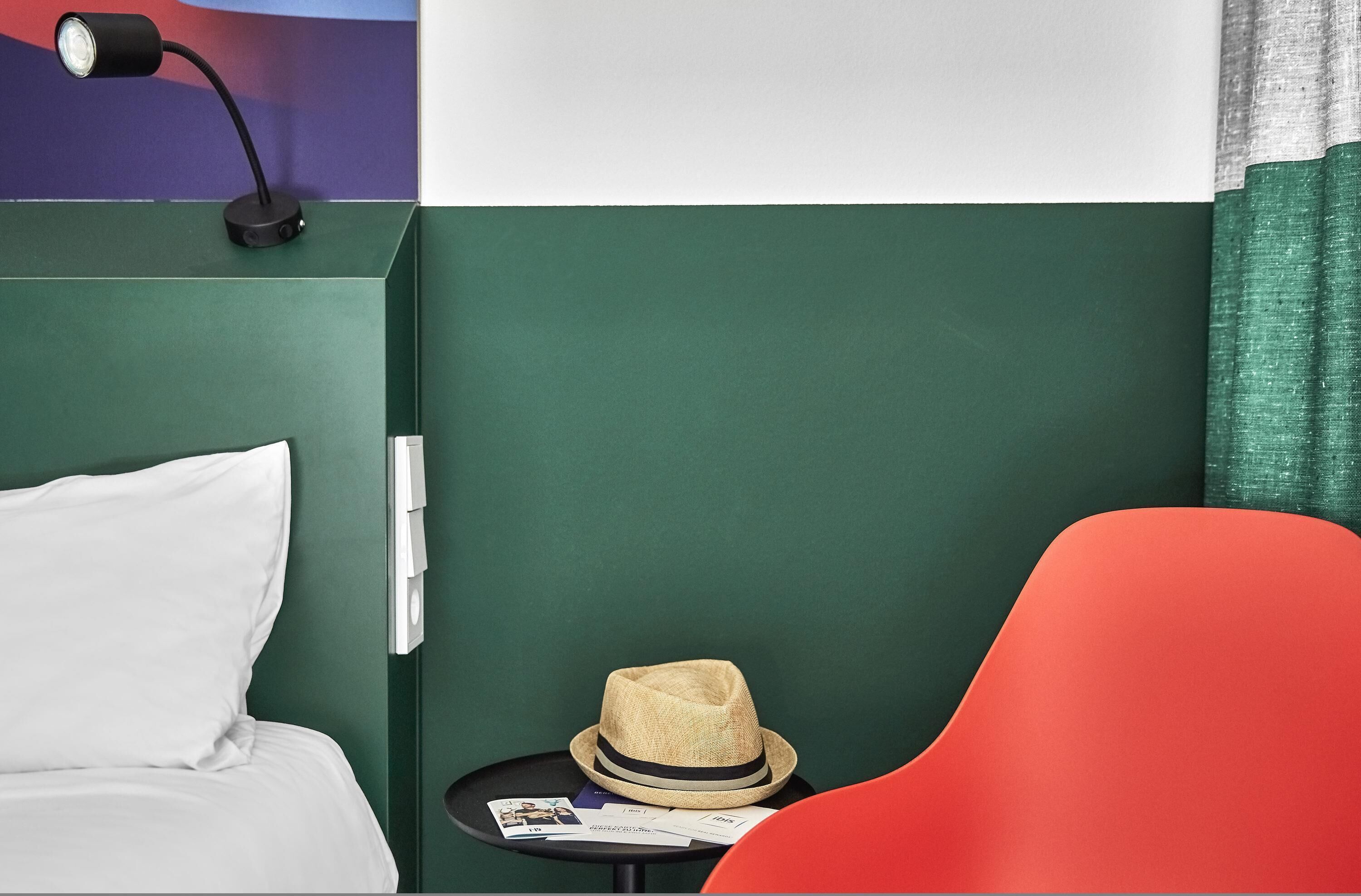A hotel room with a bed, a nightstand, a hat, and a red chair.