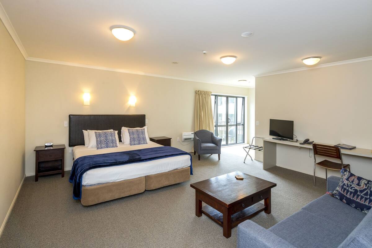 A hotel room with a large bed, a desk with a TV, a sofa, and a small table.