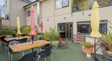 Un patio al aire libre con mesas y sillas de madera, sombrillas amarillas y rosadas, y plantas en macetas.