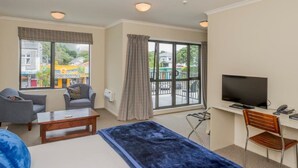 A hotel room with a bed, a desk with a TV, two armchairs, a coffee table, and large windows offering a view of the street. - Finns (Paekakariki)