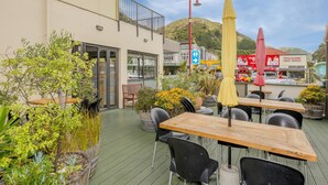 An outdoor seating area with tables and chairs, surrounded by potted plants and a view of a street with shops and a hill in the background. - Finns (Paekakariki)