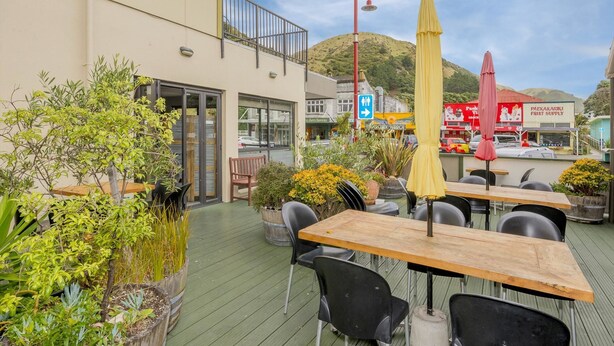 An outdoor seating area with tables and chairs, surrounded by potted plants and a view of a street with shops and a hill in the background. - Finns (Paekakariki)