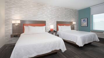 A hotel room with two beds, each with a wooden headboard and white bedding. There are bedside tables with lamps, a large window with a view, and a textured wall.