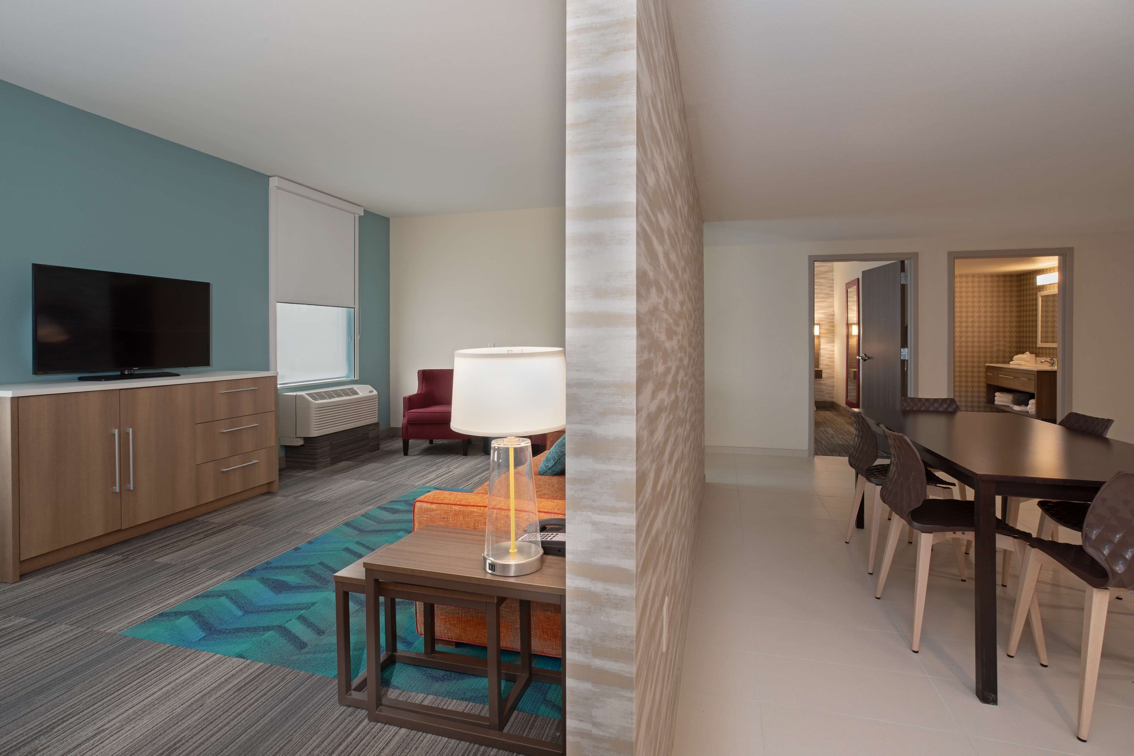 A hotel room with a TV, a sofa, a lamp, a dining table with chairs, and a bathroom visible through an open door.